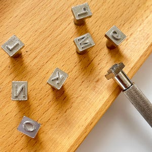 May include: Close-up of metal letter stamps arranged on a wooden surface. The stamps are in various letters, including J, L, M, K, N, and O. A metal handle tool is also visible, suggesting a crafting or personalization theme.