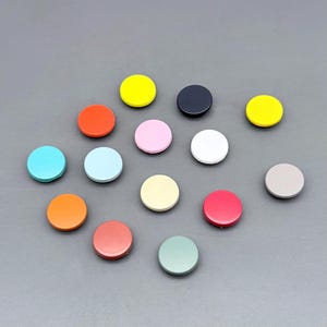 Könnte beinhalten: Eine Sammlung bunter, runder Knöpfe in verschiedenen Farbtönen, darunter Gelb, Orange, Rosa und Blau. Die Knöpfe sind auf einer grauen Oberfläche angeordnet und zeigen ihre vielfältigen Farbtöne und ihr glänzendes Finish. Diese Knöpfe könnten für Bastel- oder Nähprojekte verwendet werden.