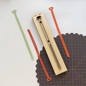 Può includere: Vista dall'alto di una fustella in legno con un taglio lungo e stretto, accompagnata da cinturini in pelle verdi e arancioni. La fustella ha il testo "SA-088 23x20". I cinturini hanno un anello a un'estremità.