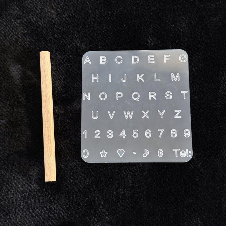 May include: A clear, square alphabet and number stamp set with a wooden handle. The stamp set includes the letters A through Z, numbers 0-9, and several symbols. The text "Tel:" is also visible on the stamp.