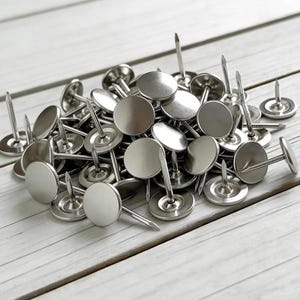 May include: A pile of silver upholstery tacks with round, flat heads and sharp, pointed nails. The tacks are scattered on a light-colored wooden surface, creating a textured and reflective display. These tacks are ideal for various craft and home decor projects.