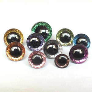 May include: A collection of colorful, round craft eyes with black pupils and glittery rims. The eyes vary in size and color, including gold, green, purple, and blue, reflecting light.