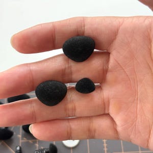 Peut inclure: Gros plan sur trois petits objets noirs en forme de galets, tenus dans une main. Les objets ont une surface texturée et varient légèrement en taille. D'autres objets similaires sont visibles en arrière-plan.