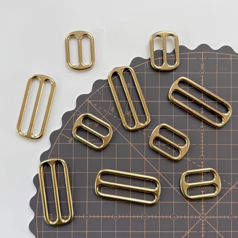 May include: An assortment of brass-coloured metal slides in different shapes and sizes. The slides are rectangular or oval with two parallel bars. These hardware pieces are used in bag making, sewing, and other craft projects.