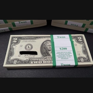 Pode incluir: Uma pilha de notas de dois dólares americanos, amarradas com faixas de papel verde. As notas apresentam um retrato de Thomas Jefferson e o texto "Two Dollars". A faixa diz "Twos" e "$200".