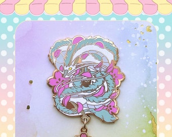 May include: A gold-tone enamel pin featuring a white dragon with blue and green scales, surrounded by pink cherry blossoms. The dragon is curled up with its head facing the viewer. The pin has a dangling charm of a pink cherry blossom.