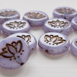May include: A collection of ten round, lavender glass beads with a gold lotus flower design. The beads are slightly textured and have a subtle shimmer.