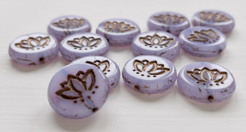 May include: A collection of ten round, lavender glass beads with a gold lotus flower design. The beads are slightly textured and have a subtle shimmer.