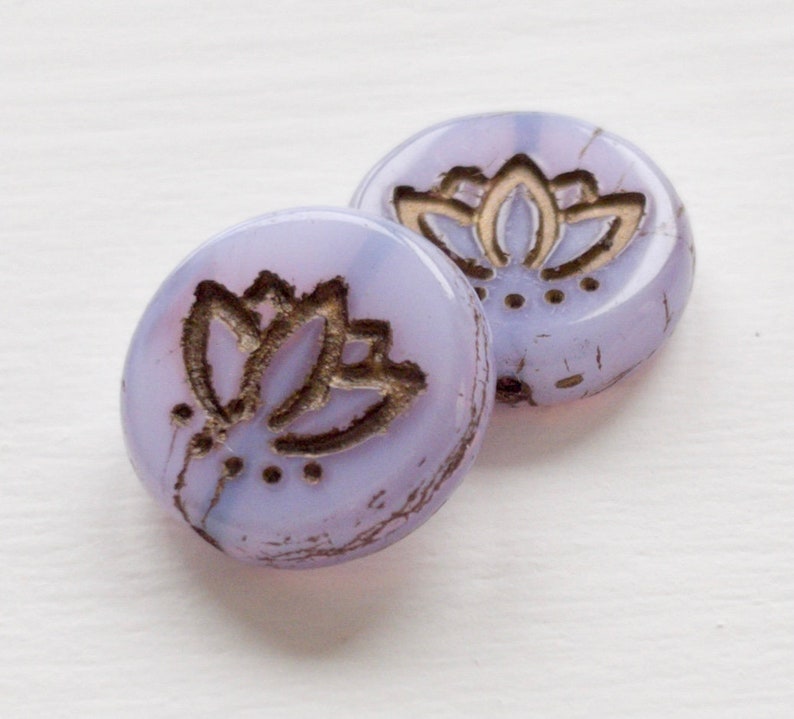 May include: Two round, lavender glass beads with a gold lotus flower design. The beads have a slightly distressed look.