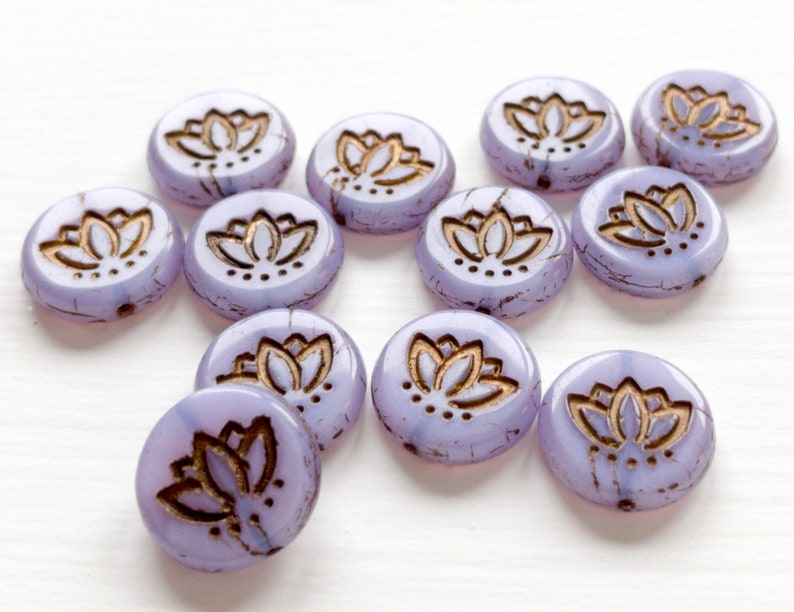 May include: A group of 12 round, lavender glass beads with a gold lotus flower design. The beads have a crackled finish.
