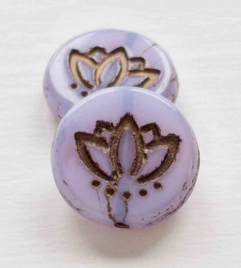 May include: Two round, lavender glass beads with a gold lotus flower design. The beads are slightly translucent and have a subtle shimmer.