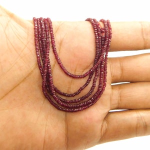 Puede incluir: Una hebra de cuentas de piedras preciosas de color rojo intenso, probablemente rubíes, enhebradas en un cordón fino. Las cuentas son facetadas y tienen una forma ligeramente irregular. La hebra está drapeada sobre una mano, mostrando la longitud y el color de las cuentas.