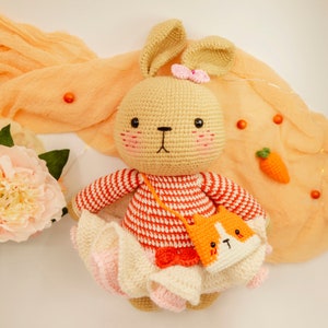 May include: A crocheted bunny rabbit toy with a pink bow, wearing a red and white striped dress and a crocheted orange and white cat purse. The bunny is sitting on a light orange fabric background.
