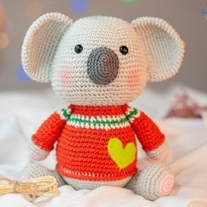 May include: A handmade crochet koala bear with a gray body, large ears, and a gray nose. The koala wears a red sweater with green and white stripes and a yellow heart. The toy is sitting on a white surface.
