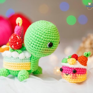 Puede incluir: Una tortuga de crochet verde que lleva un sombrero de pastel de crochet colorido con una vela rosa en la parte superior. Un pastel de crochet más pequeño con glaseado rosa y chispas de colores se encuentra junto a la tortuga.