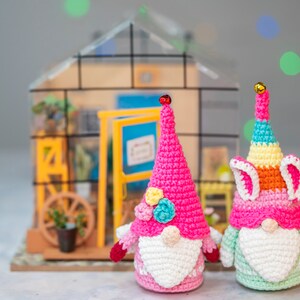 Puede incluir: Dos gnomos de crochet, uno rosa con una corona de flores y otro arcoíris con orejas de conejo, se encuentran frente a un invernadero en miniatura.