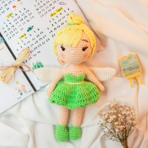 May include: A crocheted fairy doll with yellow hair, a green dress, and white wings. The doll is sitting on a white surface with a small easel and a bouquet of white flowers.