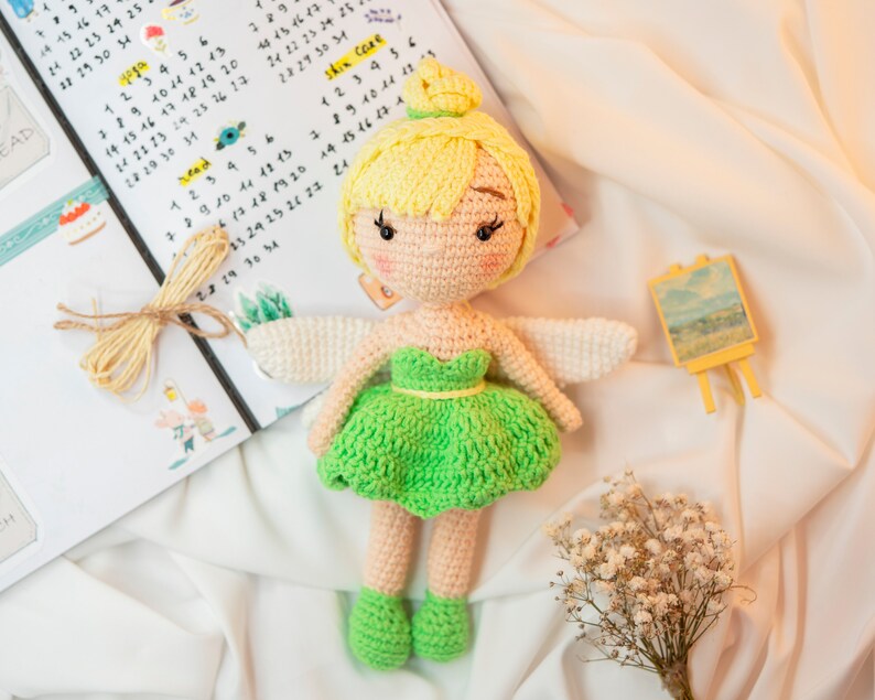 May include: A crocheted fairy doll with yellow hair, a green dress, and white wings. The doll is sitting on a white surface with a small easel and a bouquet of white flowers.