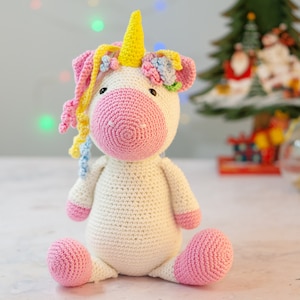 Puede incluir: Un juguete de unicornio de crochet blanco con nariz y pezuñas rosas. El unicornio tiene un cuerno amarillo y una corona de flores de colores.