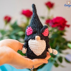 May include: A black crocheted gnome with pink heart-shaped eyes and a white heart-shaped beard. The gnome is holding a small white heart in its hand.