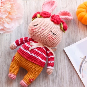 May include: A crocheted pink bunny wearing a red and white striped sweater and orange pants with a red flower crown. The bunny is lying on a wooden surface.