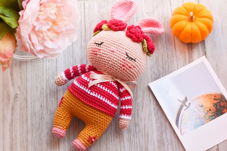 May include: A crocheted pink bunny wearing a red and white striped sweater and orange pants with a red flower crown. The bunny is lying on a wooden surface.