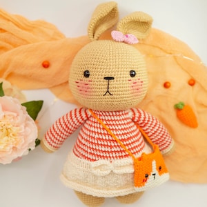May include: A crocheted bunny toy with a pink bow, wearing a red and white striped sweater and a white skirt. The bunny is holding a small orange crocheted purse with a cat face.