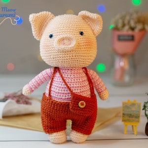 May include: A crocheted pig amigurumi toy wearing a pink and white striped shirt and brown overalls with a pocket and a button. The toy is standing on a white surface.