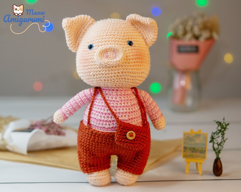 May include: A crocheted pig amigurumi toy wearing a pink and white striped shirt and brown overalls with a pocket and a button. The toy is standing on a white surface.