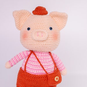 May include: A crocheted pig wearing a pink and white striped shirt and orange overalls with a small orange hat. The pig has blue eyes and a pink nose.