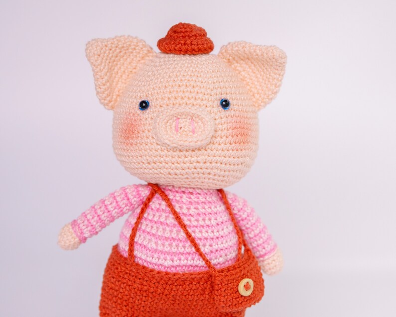 May include: A crocheted pig wearing a pink and white striped shirt and orange overalls with a small orange hat. The pig has blue eyes and a pink nose.