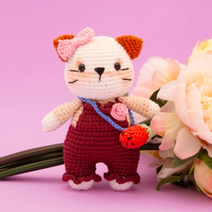May include: A crocheted cat toy wearing a red overall dress with a pink flower and a strawberry-shaped purse. The cat has a pink bow on its head and white fur with brown ears.