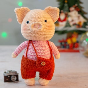 May include: A crocheted pig toy wearing a pink and white striped shirt and orange overalls. The toy has a button on the front of the overalls.