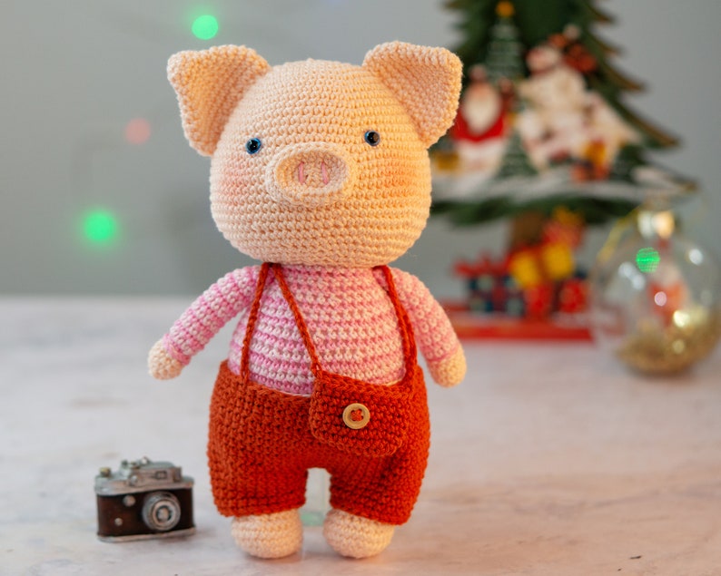 May include: A crocheted pig toy wearing a pink and white striped shirt and orange overalls. The toy has a button on the front of the overalls.