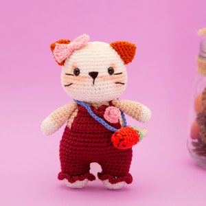 May include: A crocheted cat toy with a pink bow, brown ears, and a red overall. The cat is holding a small red strawberry.