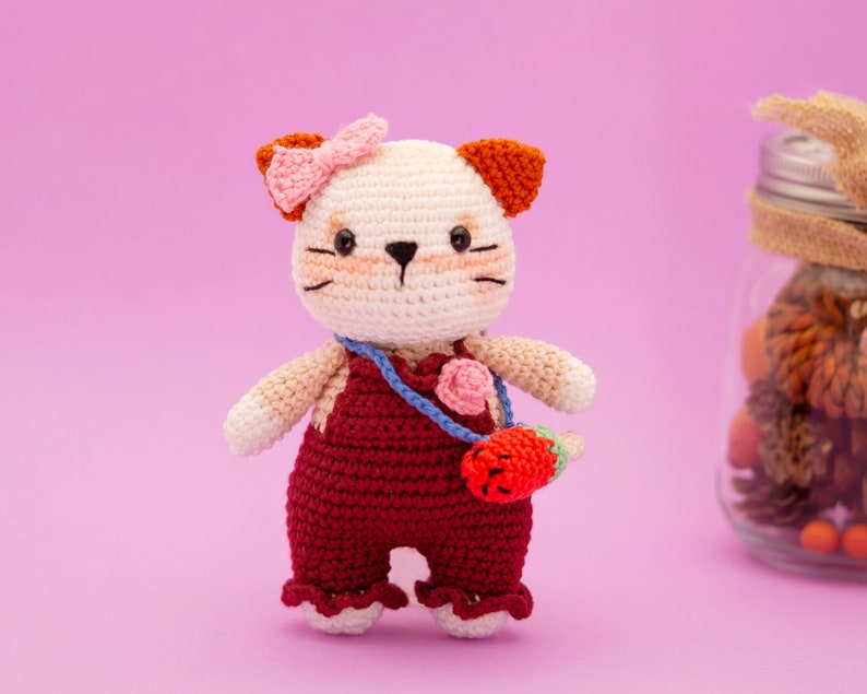 May include: A crocheted cat toy with a pink bow, brown ears, and a red overall. The cat is holding a small red strawberry.