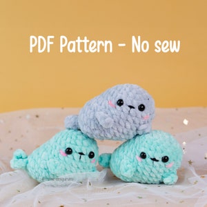 May include: Three crocheted seal plushies, one gray and two light blue, stacked on top of each other. The plushies have black eyes, pink cheeks, and a black nose. The image includes the text "PDF Pattern - No sew" at the top.