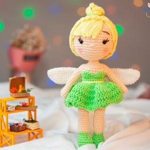 May include: A crocheted fairy doll with yellow hair, green dress, and white wings. The doll is standing on a white surface.