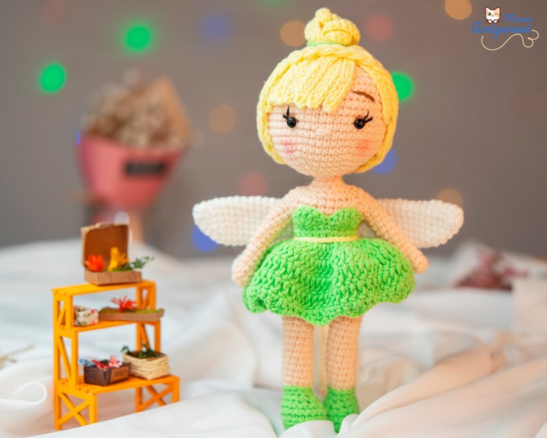 May include: A crocheted fairy doll with yellow hair, green dress, and white wings. The doll is standing on a white surface.