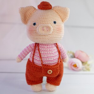 May include: A crocheted pig toy wearing a pink and white striped shirt and brown overalls with a button. The pig has a red hat and blue eyes.