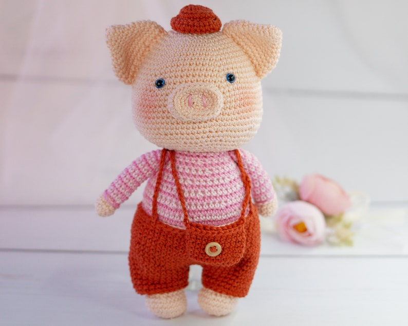 May include: A crocheted pig toy wearing a pink and white striped shirt and brown overalls with a button. The pig has a red hat and blue eyes.