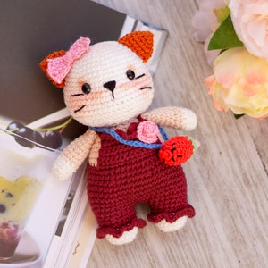 May include: A crocheted cat toy with orange ears, a pink bow, and a red overall. The cat is holding a small crocheted strawberry.