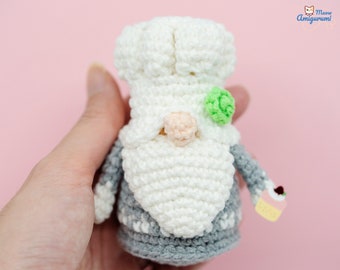 May include: A white crocheted gnome with a gray and white patterned hat and a gray body. The gnome has a white beard and a pink nose. The gnome is holding a small crocheted cupcake with a pink frosting and a brown chocolate sprinkle.