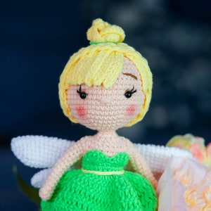 May include: A crocheted fairy doll with yellow hair, a green dress, and white wings. The doll is sitting on a pink flower.