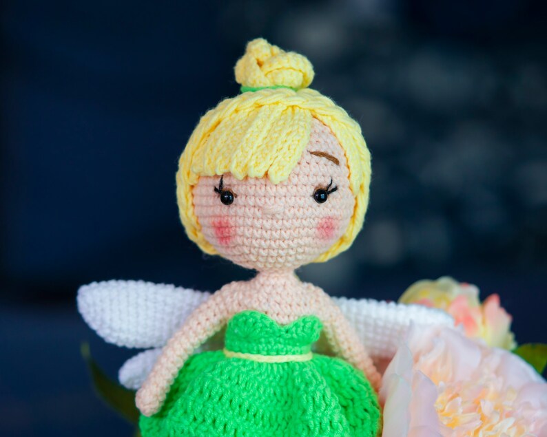 May include: A crocheted fairy doll with yellow hair, a green dress, and white wings. The doll is sitting on a pink flower.