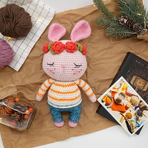 May include: A pink crocheted bunny with a flower crown, wearing a striped shirt and blue jeans. The bunny has closed eyes and a sweet expression.