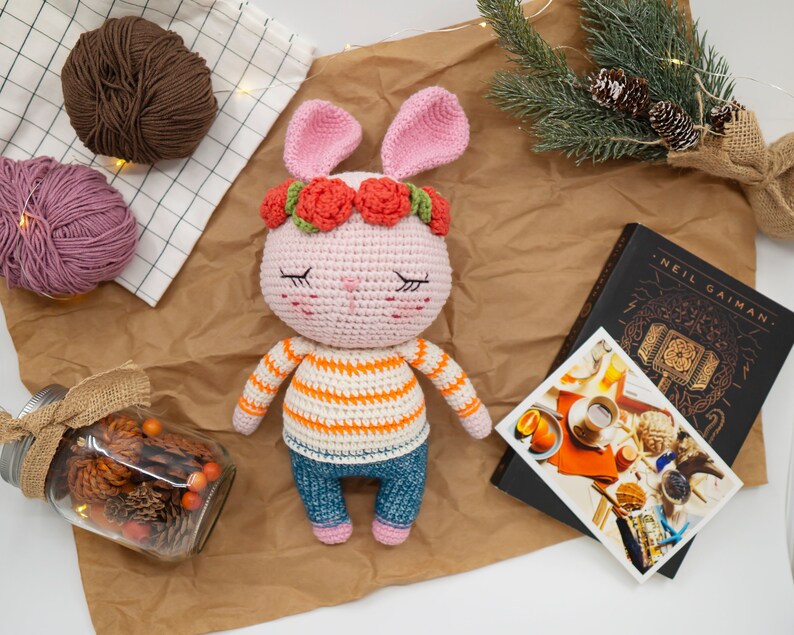 May include: A pink crocheted bunny with a flower crown, wearing a striped shirt and blue jeans. The bunny has closed eyes and a sweet expression.