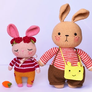 May include: Two crocheted bunny plushies, one pink with a red flower crown and a red and white striped shirt, the other brown with a red and white striped shirt and a yellow bag.