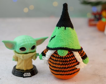 May include: A green and black crocheted gnome with a white beard and orange and black striped body. The gnome is wearing a black witch hat. A green and yellow Star Wars character is standing next to the gnome.