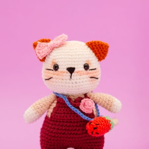 May include: A crocheted cat toy with a pink bow, white fur, red overalls, and a blue strap with a pink flower and a red strawberry. The toy is against a pink background.